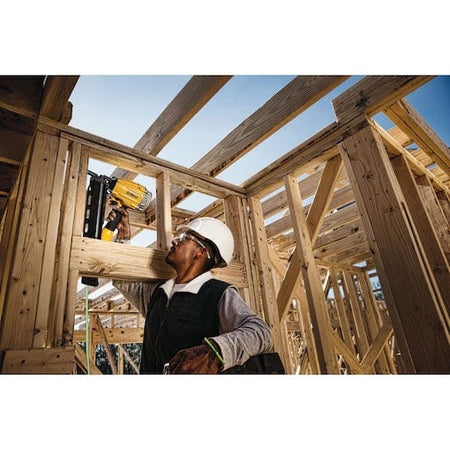 Construction worker using yellow and black DEWALT nail gun to secure wooden framing, wearing safety gear at an active jobsite under clear sky.