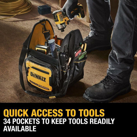 Lifestyle image of a person holding a DEWALT tool tote bag filled with hand tools in a construction setting. The black and yellow bag displays “WATER RESISTANT CONSTRUCTION” text and organized outer pockets. The person stands on a dusty floor with wood and building materials in the background.