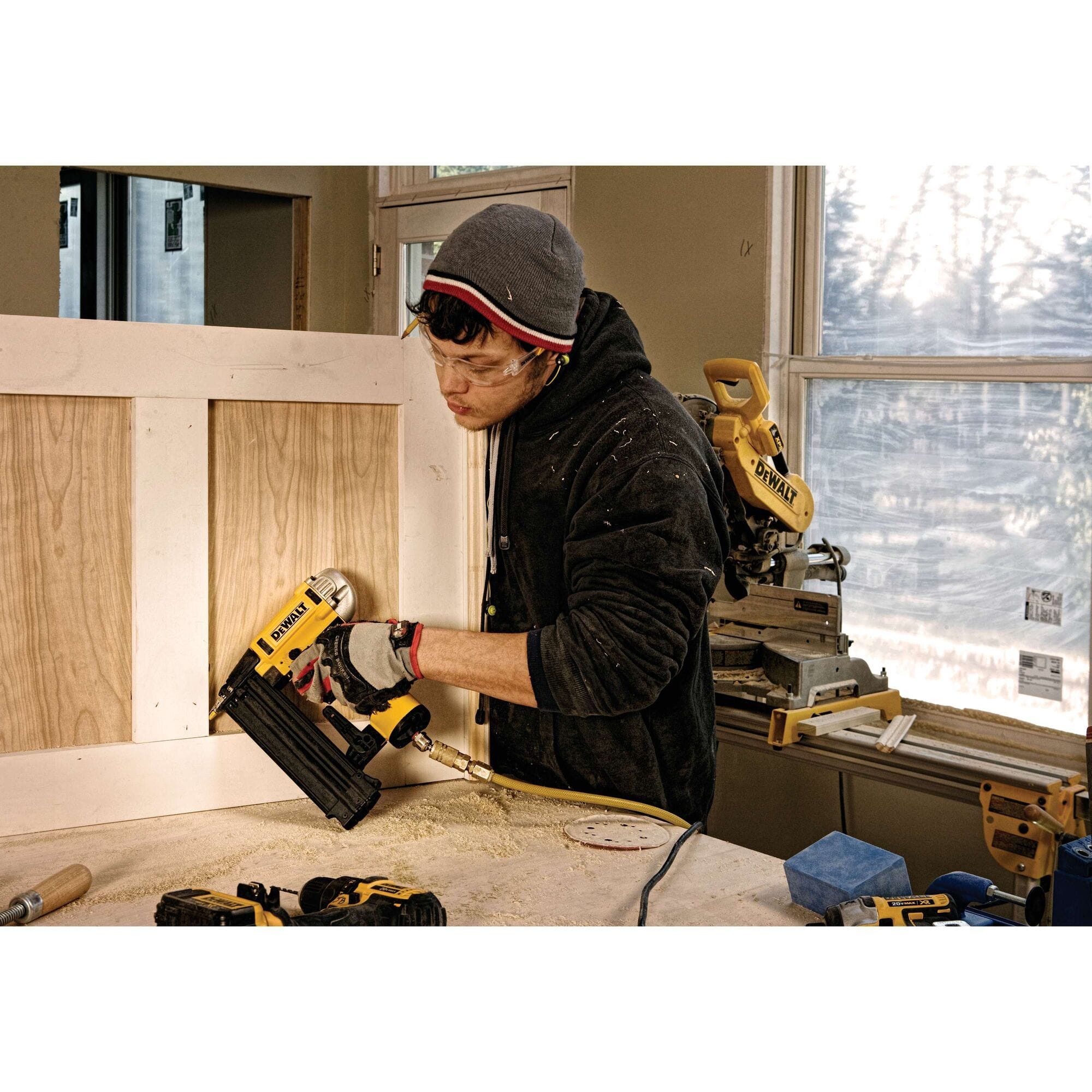 Wide shot of a woodworker in a workshop using the DEWALT 18 gauge pneumatic finish nailer to assemble a wood panel, with various DEWALT power tools in the background.
