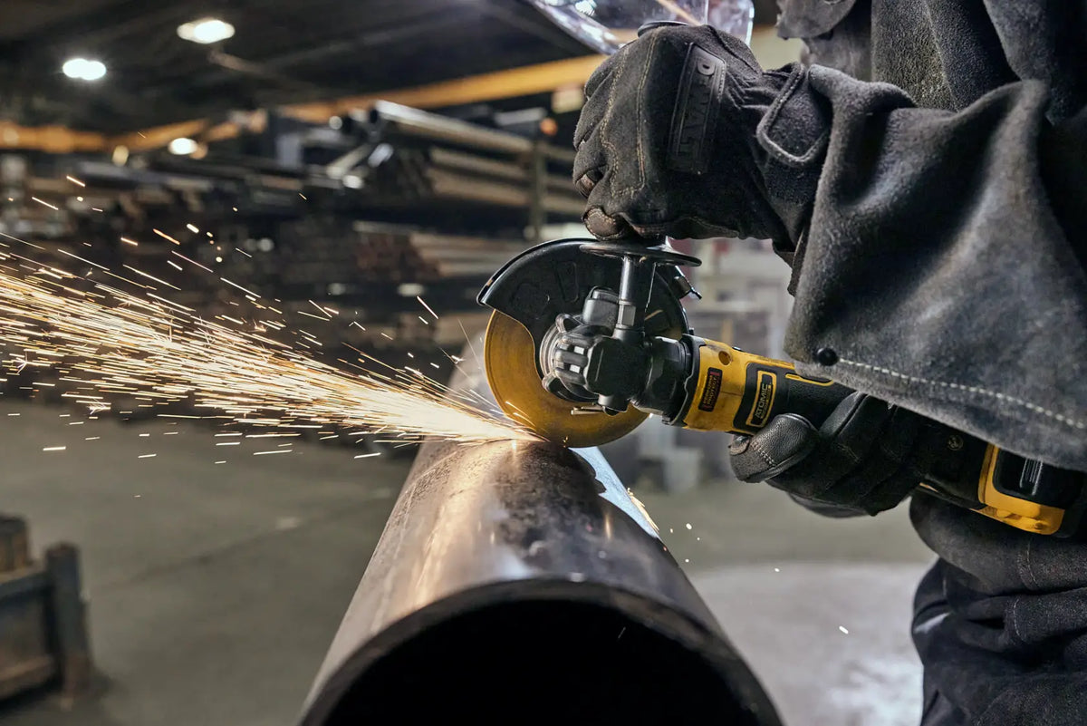 An action lifestyle shot of a professional welder using the DEWALT ATOMIC 20V MAX cordless angle grinder to grind a large metal pipe. The image captures a shower of sparks, demonstrating the tool's 11,000 RPM power and the effective protection of the adjustable safety guard.