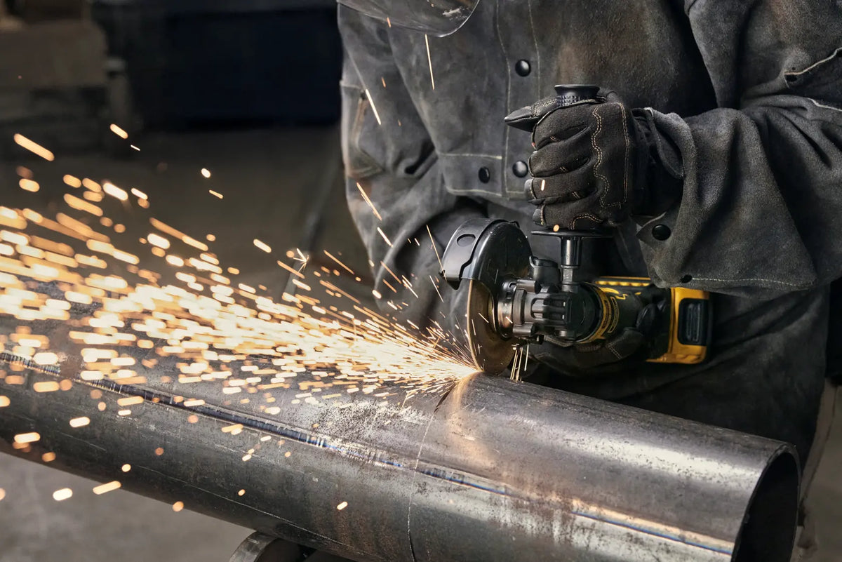 An action lifestyle shot of a professional tradesperson using the DEWALT 20V MAX ATOMIC cordless angle grinder to cut a thick metal pipe. The image shows the grinder in a horizontal position with sparks flying, highlighting the high-speed 11,000 RPM performance and the protective safety guard in use.