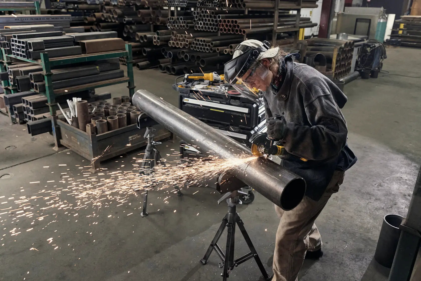 A wide action shot of a professional wearing a safety face shield and gloves while using the DEWALT ATOMIC 4-1/2 inch angle grinder on a long metal pipe in a fabrication shop. The image emphasizes the tool's portability and ease of use in industrial environments for precise metal cutting and grinding.