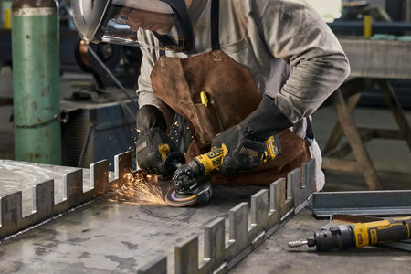 A close-up action shot of a professional fabricator wearing a leather apron and safety gear using the DEWALT DCG402B cordless grinder to finish a series of metal notches. The sparks illustrate the brushless motor power, while the compact ATOMIC series design is shown accessing tight areas on a large steel workpiece.