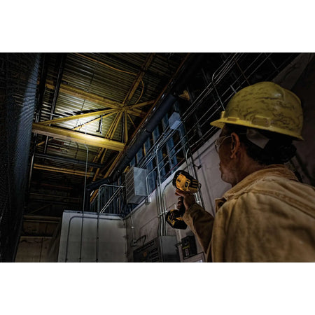 An action lifestyle shot from the perspective of a technician holding the DEWALT 20V MAX spotlight. The high-intensity beam is shown illuminating a complex ceiling structure in a dark industrial facility, demonstrating the light's long-range spotting capability.