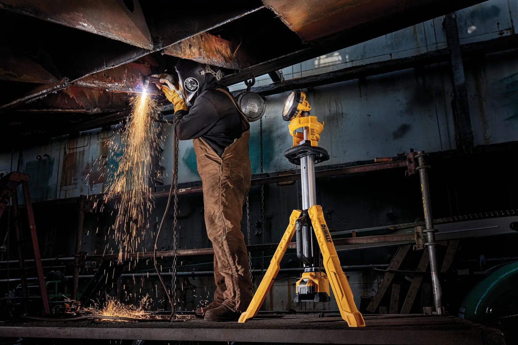 A wide action shot showing the DEWALT 20V MAX tripod light illuminating a fabrication shop. The light is positioned to provide clear visibility for a technician performing heavy-duty metal work, demonstrating the IP55 water resistance and stability of the stand.
