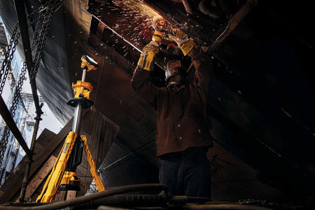 A perspective action shot of a professional tradesman using a grinder under the illumination of the DEWALT cordless tripod light. The image emphasizes the natural white light output that provides precise visibility for dangerous tasks in dimly lit outdoor or indoor areas.