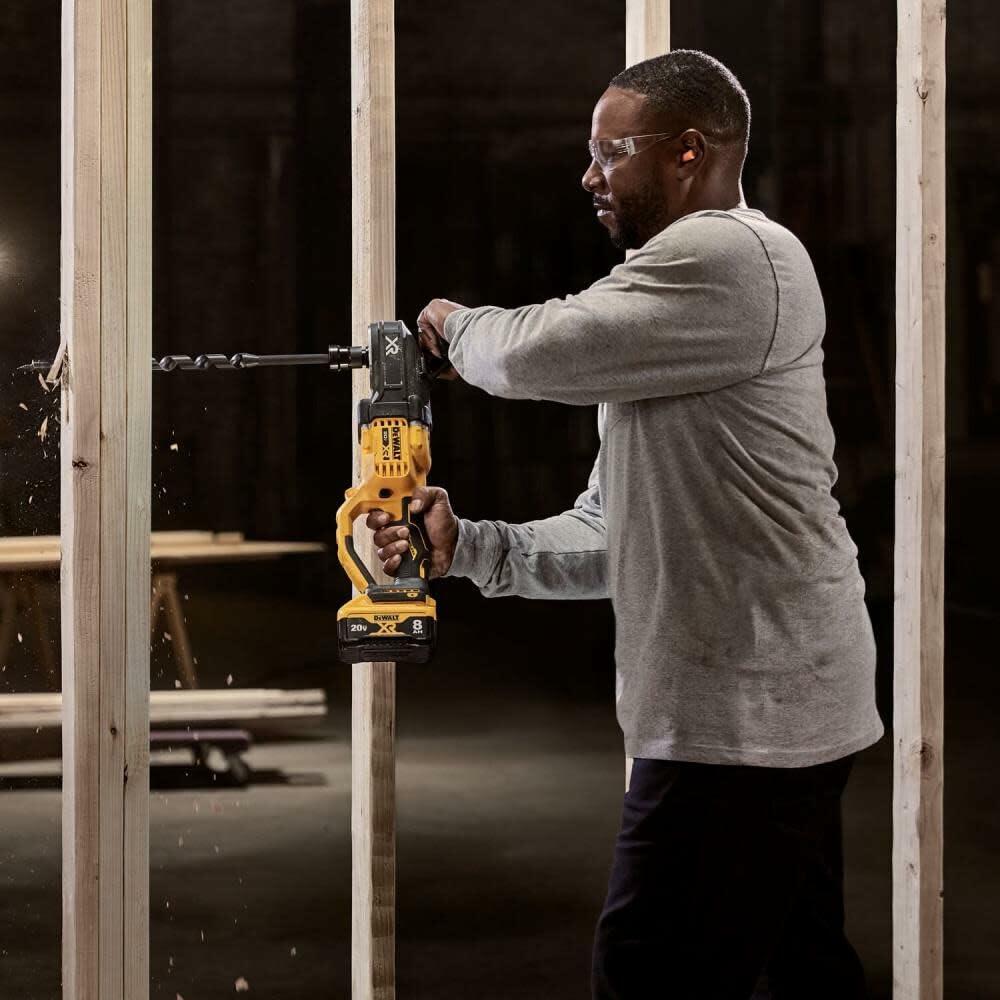 A tradesperson wearing safety gear uses the DEWALT Stud and Joist Drill to bore a large hole horizontally through a wooden stud in a wall frame.