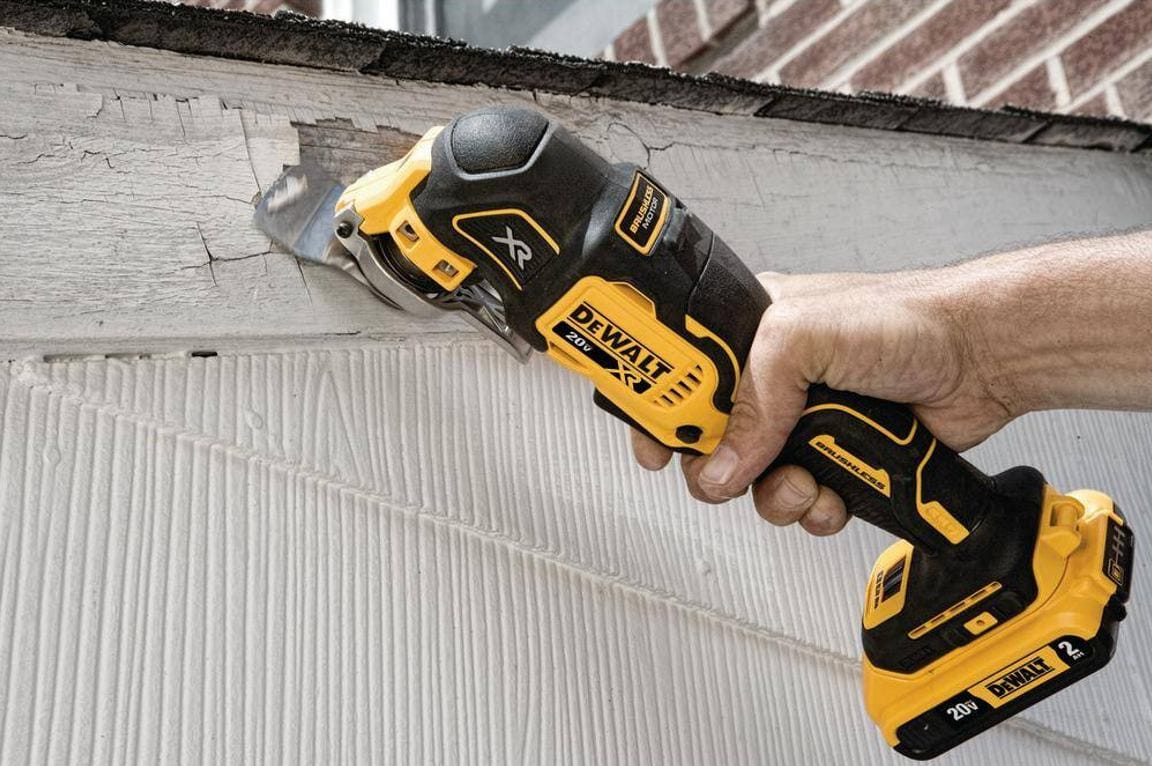A close-up application photo showing a user operating the DEWALT DCS356 oscillating tool to scrape away old paint and damaged wood from a house exterior, highlighting the brushless motor's power for restoration tasks.