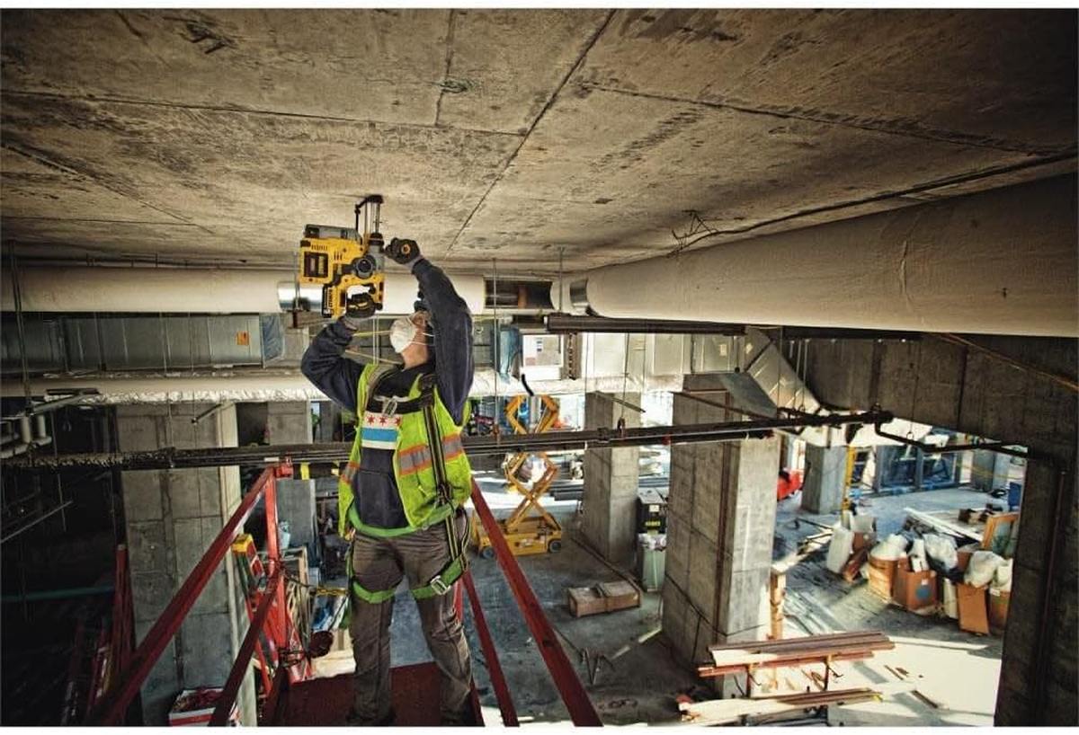 An electrician using the DEWALT 20V MAX XR cordless rotary hammer to install conduit clamps on a concrete ceiling at a large-scale industrial construction site.