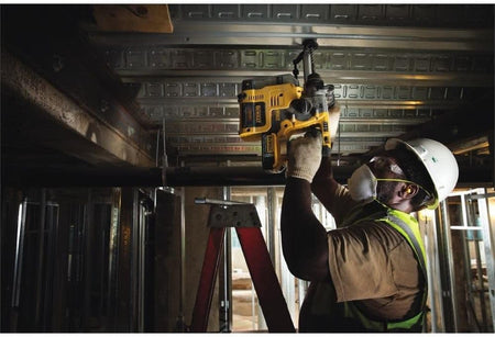 Construction worker on a ladder using the compact DEWALT SDS-Plus rotary hammer to drill into metal decking for overhead mechanical and electrical installations.