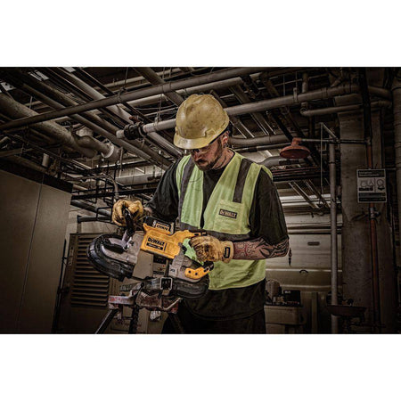 An action lifestyle shot of a mechanical contractor wearing a hard hat and safety vest while using the DEWALT 20V MAX XR portable band saw. The technician is performing an overhead cut on industrial piping in a dimly lit mechanical room, showcasing the tool's cordless portability.
