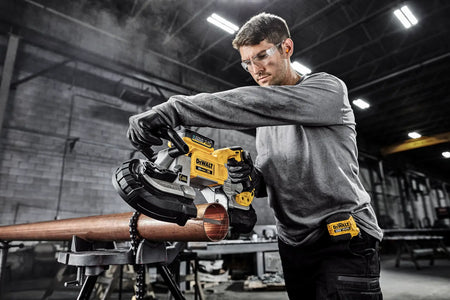 A wide lifestyle shot of a professional tradesman using the DEWALT deep cut band saw to slice through a large diameter copper pipe. The image showcases the tool's smooth operation and the user's ergonomic stance while working in a large shop environment.