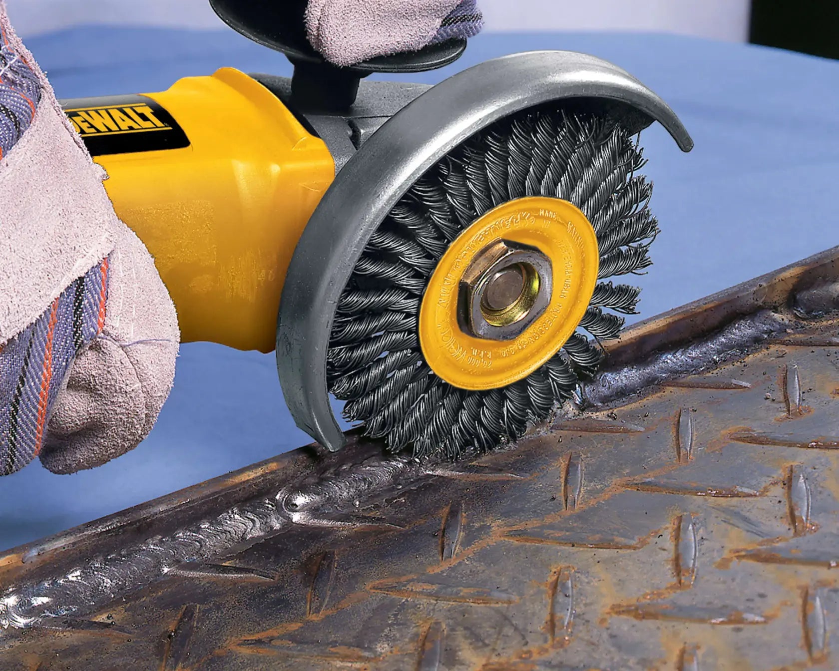 A close-up action shot of the DEWALT stringer bead wire wheel attached to an angle grinder. The tool is being used by a worker in protective gloves to clean a weld bead on a piece of textured diamond plate metal, demonstrating effective surface preparation.