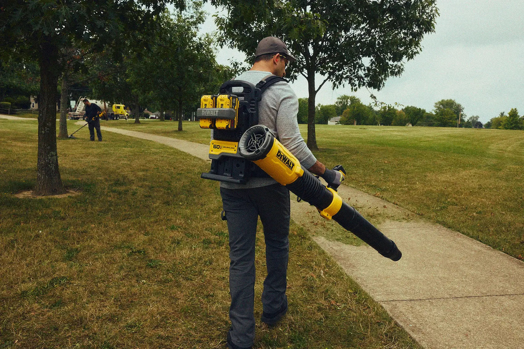 Groundskeeper operating the DEWALT 60V MAX FLEXVOLT Brushless Backpack Blower DCBL570Z2 with dual battery packs during large‑area landscaping cleanup in a park setting.