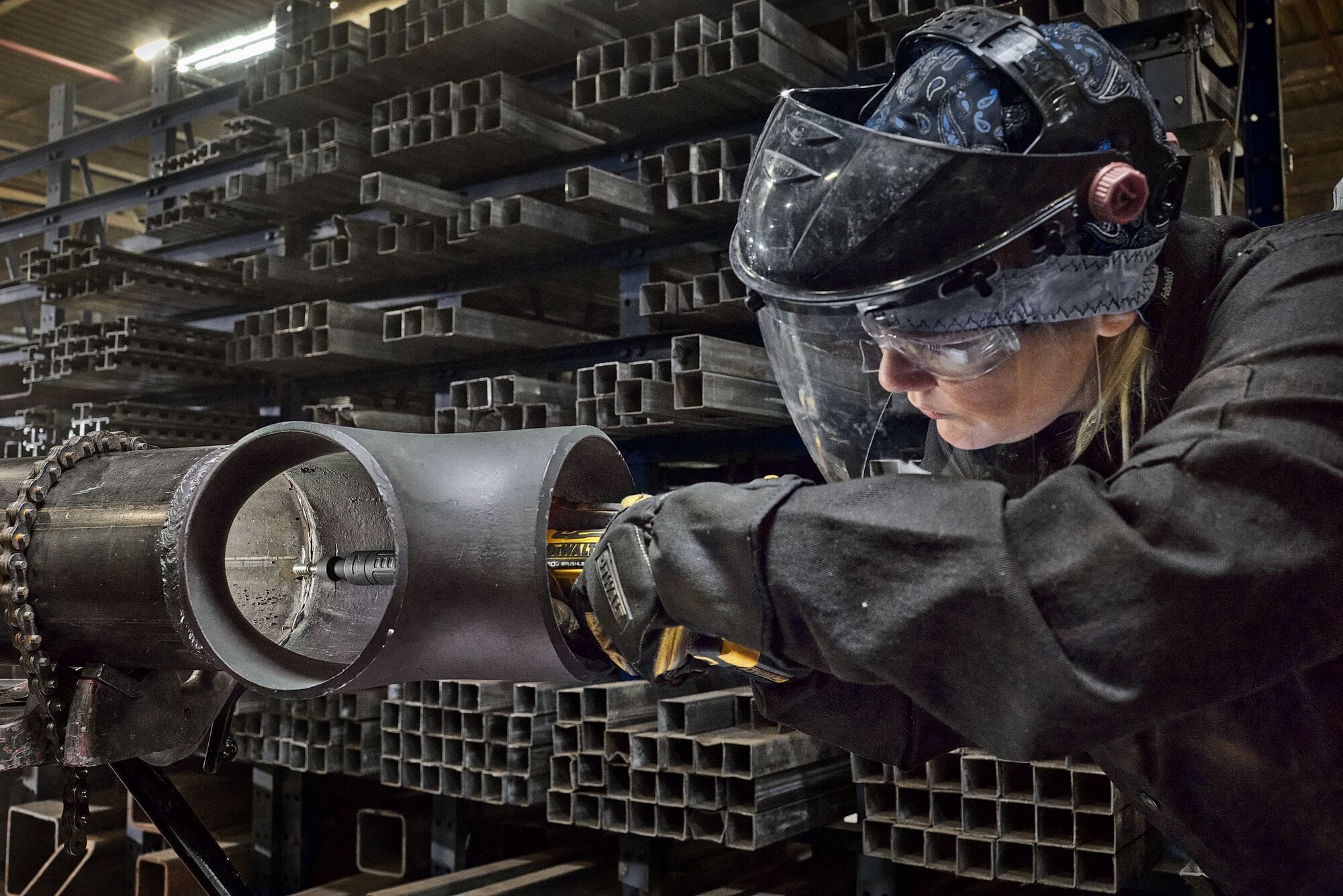 Professional metalworker wearing a face shield using the DEWALT ATOMIC 20V MAX extended die grinder to access and grind the inside of a large steel pipe fitting.