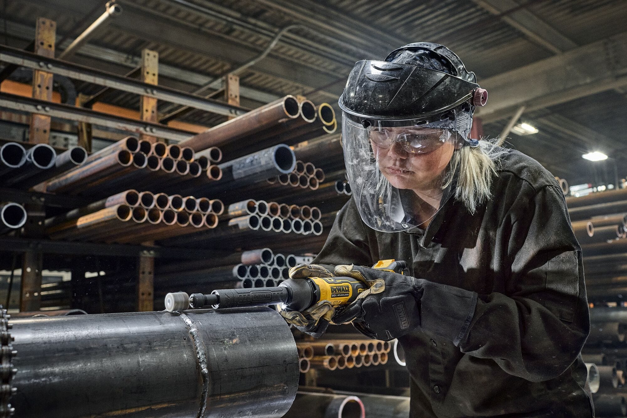 Fabricator in a metal shop using the DEWALT DCG420XGE2 20V MAX extended die grinder to polish an exterior weld on a heavy structural steel pipe.