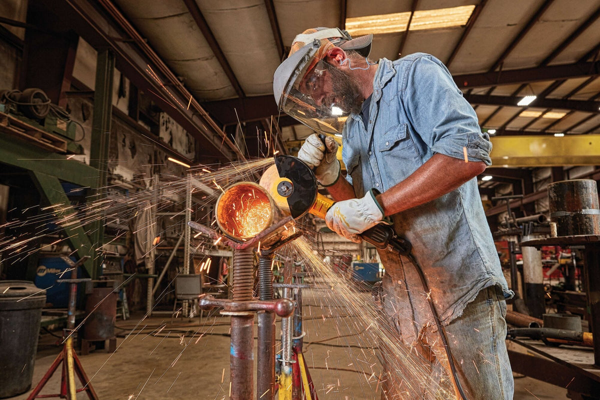 Wide angle action photograph of a fabricator in a manufacturing facility using a 5 to 6 inch DEWALT corded angle grinder tool to finish a pipe section clamped onto an adjustable screw support stand.