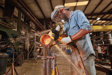 Wide angle action photograph of a fabricator in a manufacturing facility using a 5 to 6 inch DEWALT corded angle grinder tool to finish a pipe section clamped onto an adjustable screw support stand.