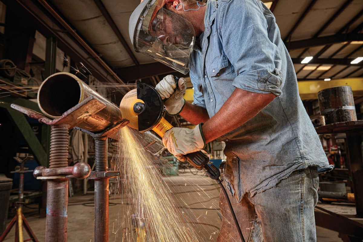 Industrial action shot of a trade professional wearing eye protection and a face shield using a DEWALT brushless angle grinder on a steel pipe workpiece while brilliant sparks spray across a workshop.