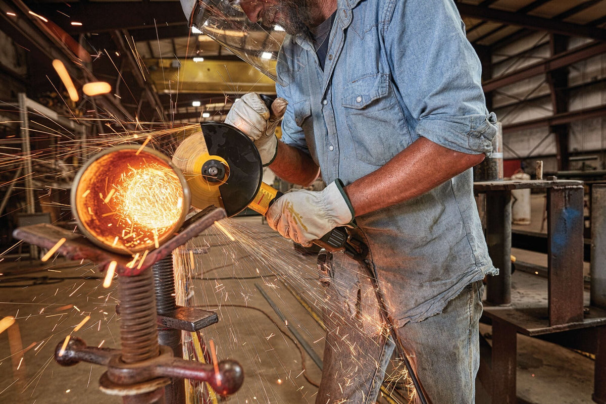 Alternative industrial perspective of a welder manually deburring and grinding down a thick cylindrical metal pipe part over a steel workbench frame creating a wide shower of sparks inside a production shop.