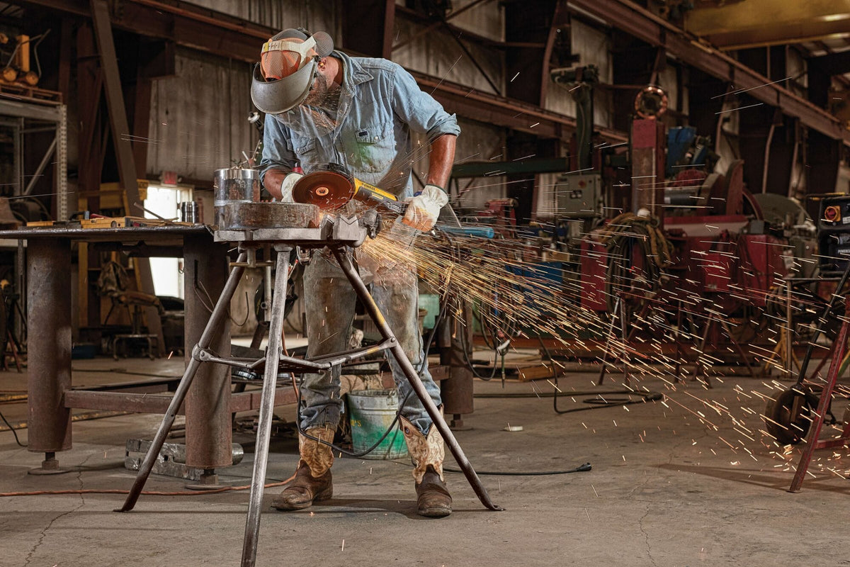 Full-length action shot of a professional welder wearing a safety mask and heavy leather gloves using a DEWALT 7 inch brushless electric corded rat tail angle grinder to remove surface welds from a thick pipe segment clamped in a metal stand.