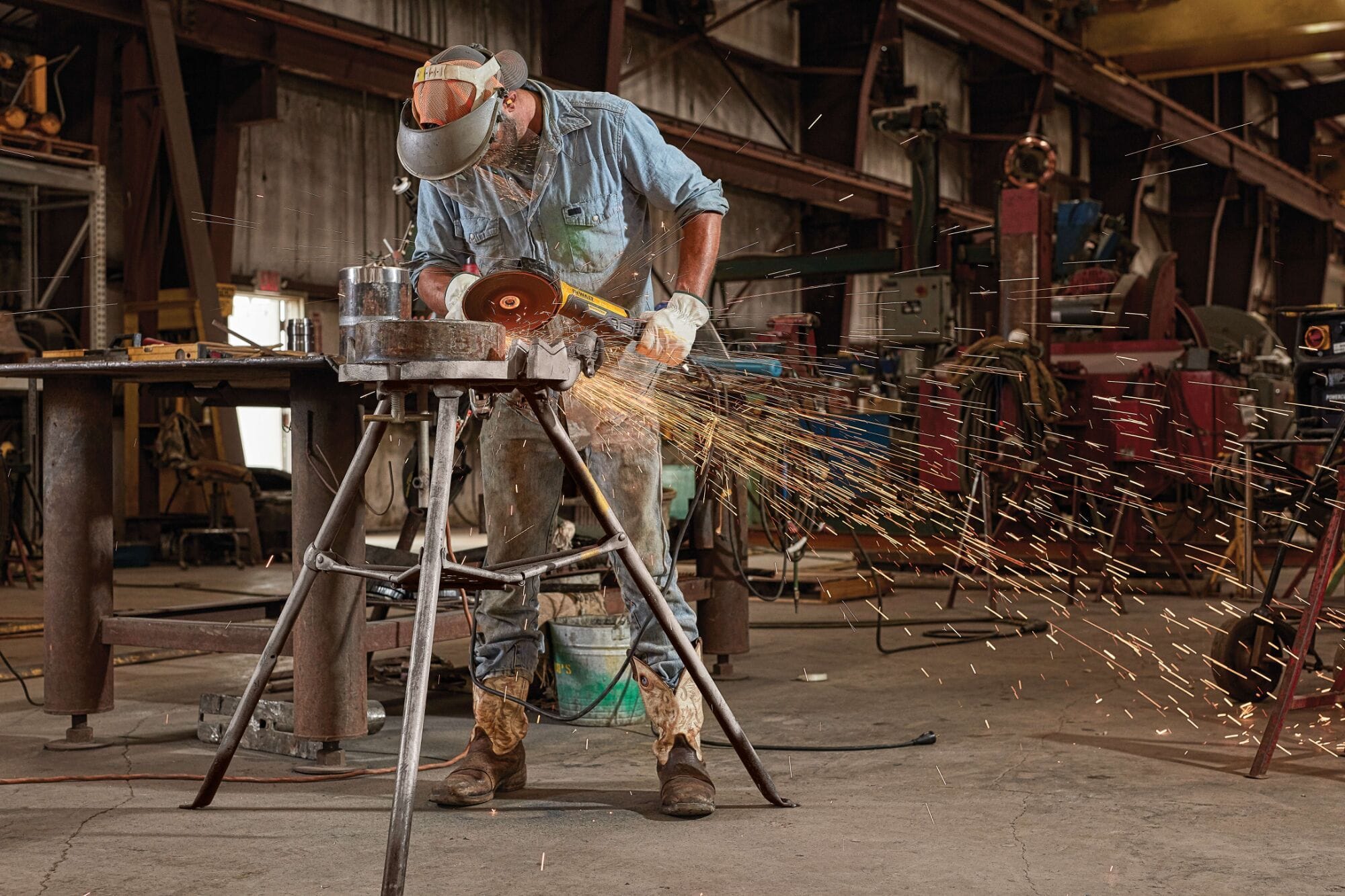 Full-length action shot of a professional welder wearing a safety mask and heavy leather gloves using a DEWALT 7 inch brushless electric corded rat tail angle grinder to remove surface welds from a thick pipe segment clamped in a metal stand.