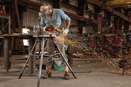 Full-length action shot of a professional welder wearing a safety mask and heavy leather gloves using a DEWALT 7 inch brushless electric corded rat tail angle grinder to remove surface welds from a thick pipe segment clamped in a metal stand.