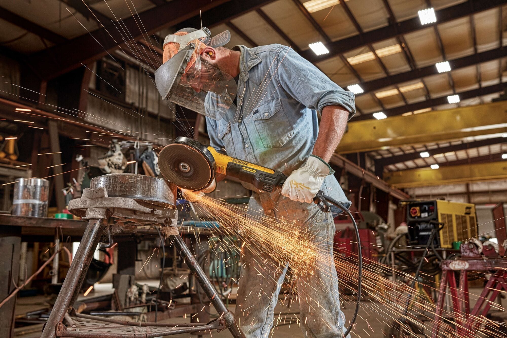 Alternative industrial perspective of a welder manually deburring and grinding down a thick cylindrical metal pipe part over an iron support framework frame creating a wide shower of sparks inside a production shop.