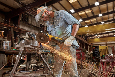 Alternative industrial perspective of a welder manually deburring and grinding down a thick cylindrical metal pipe part over an iron support framework frame creating a wide shower of sparks inside a production shop.