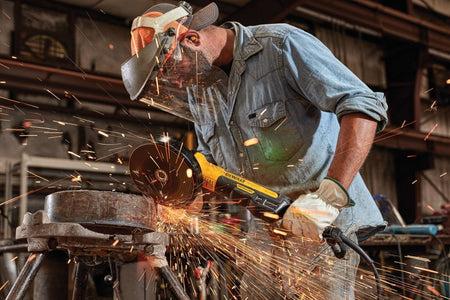 Side view perspective of an industrial worker operating a DEWALT 7 inch 8000 RPM angle grinder tool on a pipeline metal joint workpiece while a dense shower of grinding sparks shoots across a fabrication shop floor.