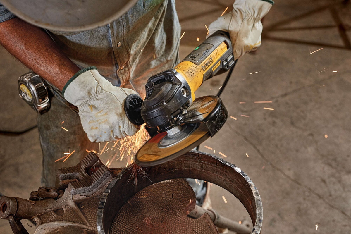 Top-down high angle macro shot of a tradesperson utilizing a DEWALT brushless paddle switch electric grinder to smooth the edges of a curved industrial metal pipe ring while wearing heavy-duty safety gloves.