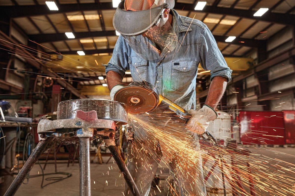 Industrial action shot of a trade professional wearing eye protection and a face shield using a DEWALT brushless angle grinder on a steel pipe workpiece while brilliant sparks spray across a workshop.