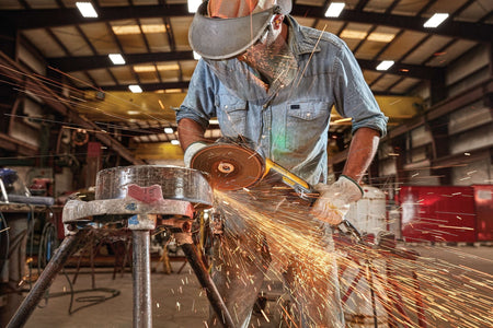 Industrial action shot of a trade professional wearing eye protection and a face shield using a DEWALT brushless angle grinder on a steel pipe workpiece while brilliant sparks spray across a workshop.