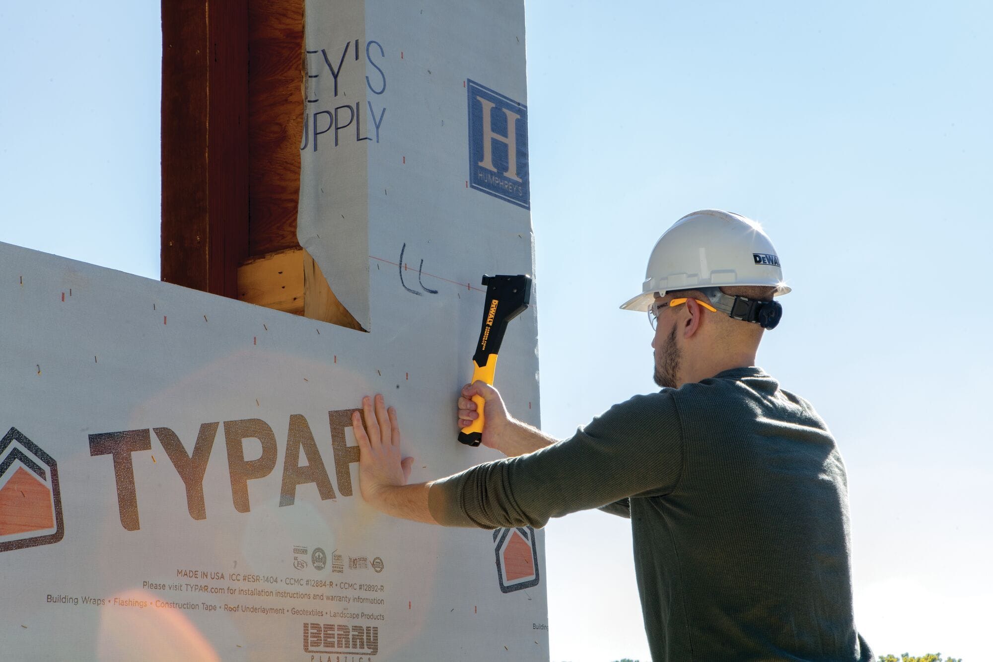 A worker wearing a hard hat and safety glasses using the DEWALT DWHT75900 hammer tacker to install TYPAR house wrap, highlighting the tool's effectiveness for vertical fastening.