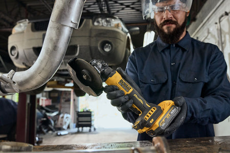 Automotive mechanic wearing safety glasses and a face shield holding the DEWALT DCG400S2 compact cordless angle grinder near a vehicle exhaust system pipe inside a repair garage workshop