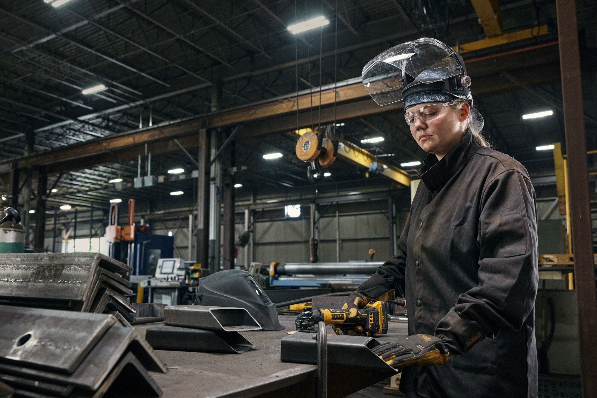 Industrial manufacturing fabricator wearing a protective face shield and safety gloves operating the DEWALT ATOMIC 20V compact cordless grinder at a steel work table inside a large metal fabrication plant