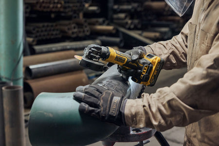 Close-up action view of a structural metalworker wearing thick leather safety gloves using the DEWALT 4 inch cordless angle grinder to prepare and grind a curved steel pipe elbow piece