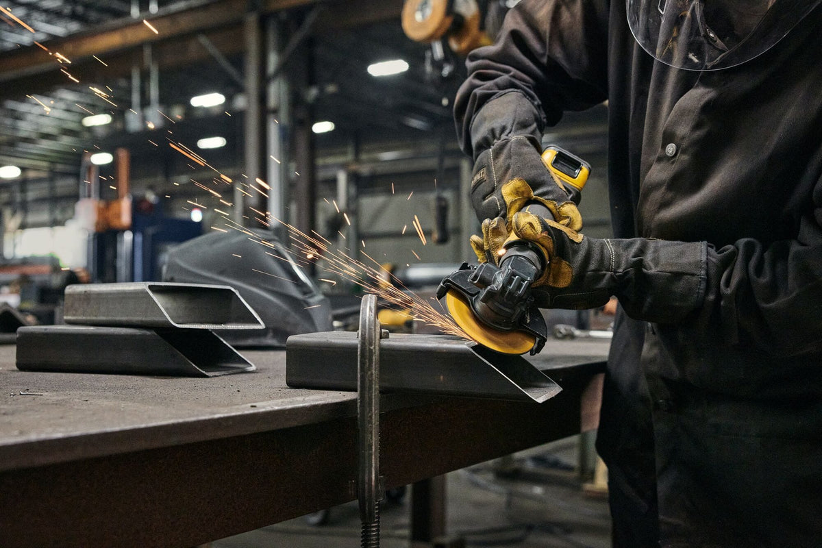 Metal fabricator using the DEWALT compact cordless grinder to cut through a structural steel box channel tube clamped to a metal workbench while throwing bright orange friction sparks