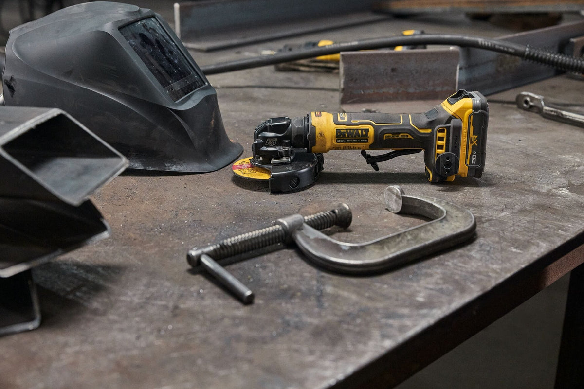 DEWALT ATOMIC 20V MAX compact cordless angle grinder resting horizontally on a metal workshop fabrication bench next to a black welding helmet hood and a heavy duty steel C-clamp tool