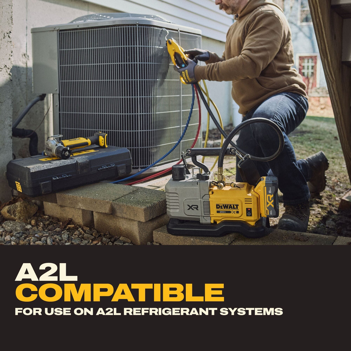 Residential AC technician kneeling outdoors near a concrete foundation wall next to an outdoor condenser unit, utilizing manifold gauges with blue and red lines connected to a DEWALT DCE510 cordless vacuum pump for A2L system evacuation.