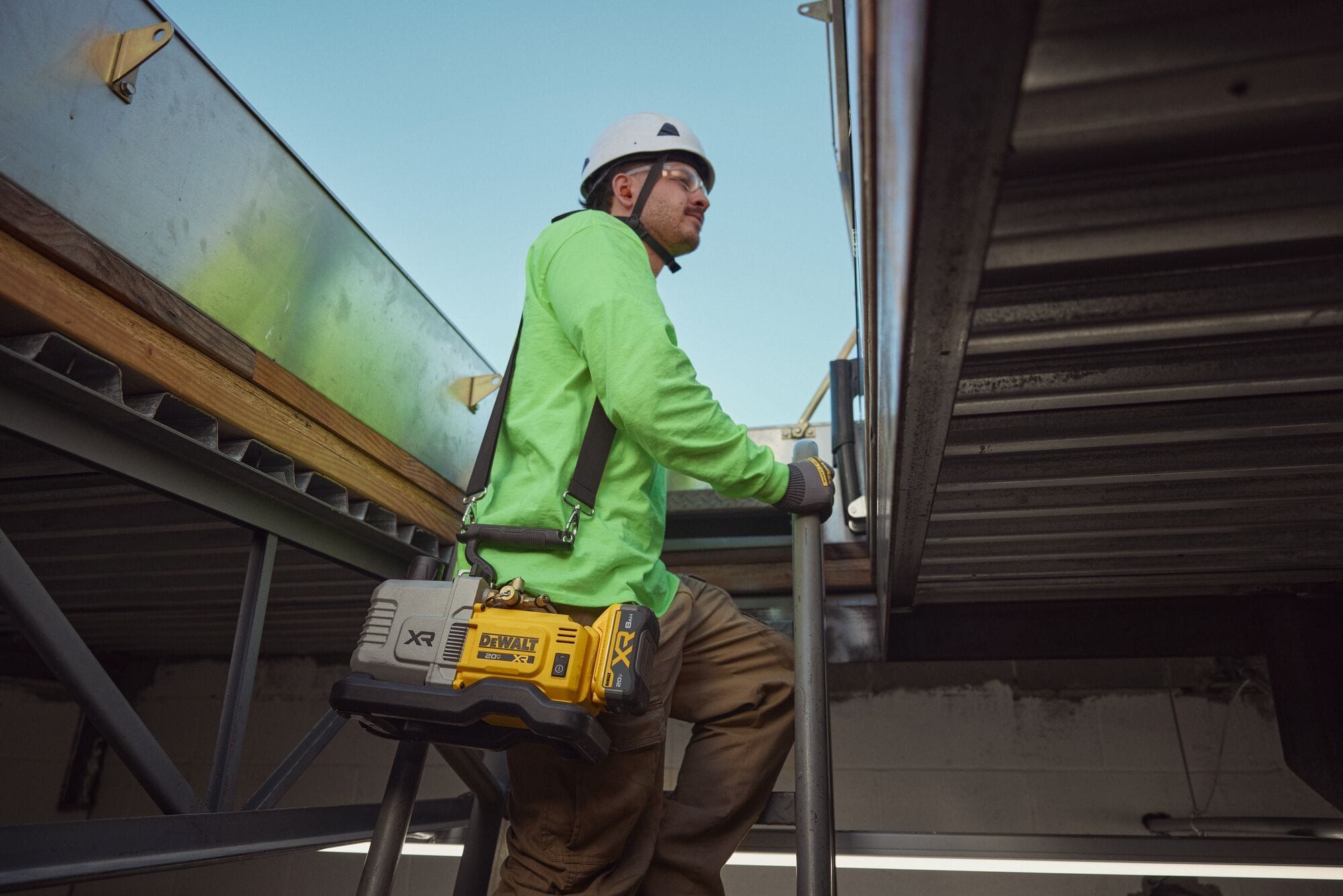 Commercial pipe trade technician with protective eyewear and a white hard hat walking up an outdoor metal building staircase with the DEWALT 20V MAX XR 5 CFM cordless vacuum pump hanging from his shoulder by the included black strap.