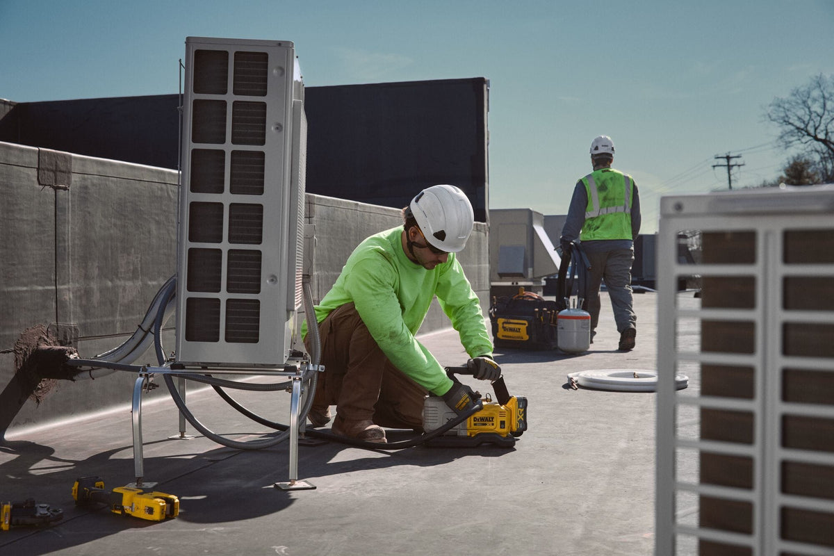 Industrial rooftop shot of an HVAC service mechanic in a green shirt kneeling next to a large commercial central air unit, checking connections with the DEWALT cordless vacuum pump resting on a flat membrane roof while another contractor walks in the background.