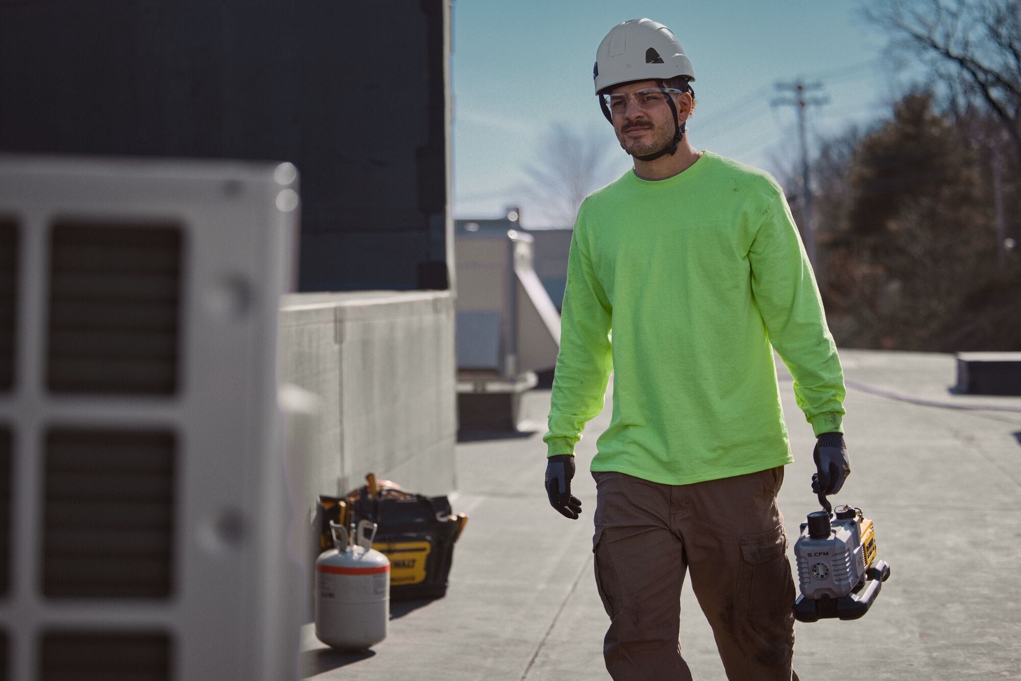 Rooftop service professional wearing safety gear walking across a flat commercial building roof while carrying the compact DEWALT 20V MAX XR 5 CFM cordless vacuum pump by its top composite handle next to HVAC condenser units.