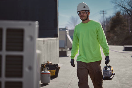 Rooftop service professional wearing safety gear walking across a flat commercial building roof while carrying the compact DEWALT 20V MAX XR 5 CFM cordless vacuum pump by its top composite handle next to HVAC condenser units.