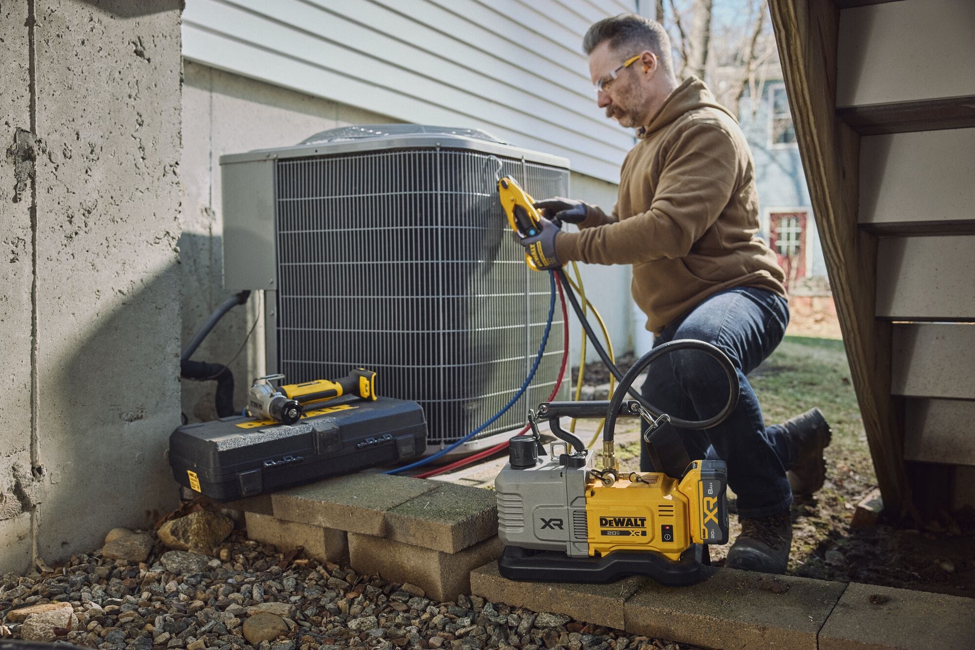 Side angle view of a service technician setting up a DEWALT DCE510 cordless vacuum pump below a flight of outdoor wooden stairs to perform a certified A2L refrigerant evacuation on a residential air conditioning condenser.