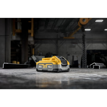A lifestyle shot of the DEWALT 20V MAX POWERSTACK 5Ah battery pack resting on a concrete floor in a workshop setting. The image highlights the battery's compact footprint and rugged design in a professional industrial environment.