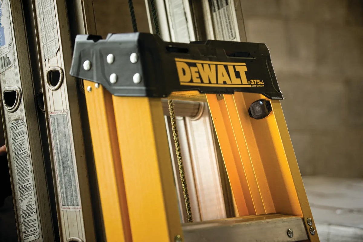 An action shot of the DEWALT DCE041 Bluetooth tag securely mounted to the side rail of a yellow fiberglass step ladder. The small, discreet profile of the tag is shown, illustrating how it can be placed inconspicuously on large jobsite assets to prevent theft and aid in inventory identification.