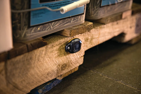 A close-up photograph of a DEWALT DCE041 tag screwed into the side of a wooden shipping pallet. The blue LED indicator light is illuminated, showcasing the tag's ability to track bulk materials and non-powered jobsite assets through the Bluetooth inventory management system.