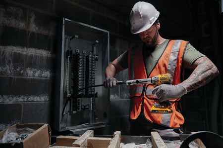 Commercial electrician wearing a hard hat and safety vest using the DEWALT DCE151B 20V MAX XR brushless cable stripper to prep heavy-gauge wire in front of a large electrical distribution panel.