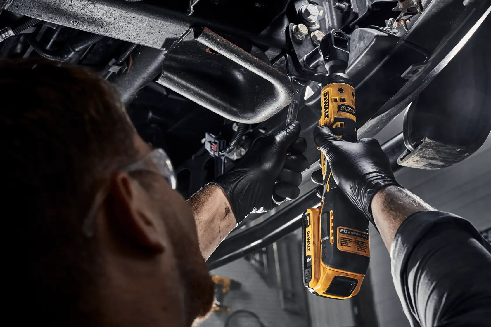 Professional mechanic utilizing the compact DEWALT 20V MAX brushless 1/2-inch cordless ratchet alongside a hand wrench to efficiently tighten suspension bolts in a tight automotive workspace.